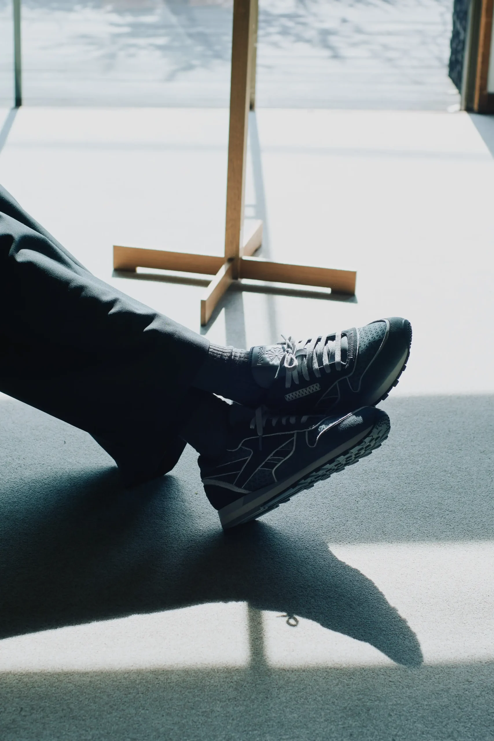 Black Reebok sneakers worn with tailored pants, shown in a sunlit indoor setting reflecting minimal, contemporary style in the global fashion market.