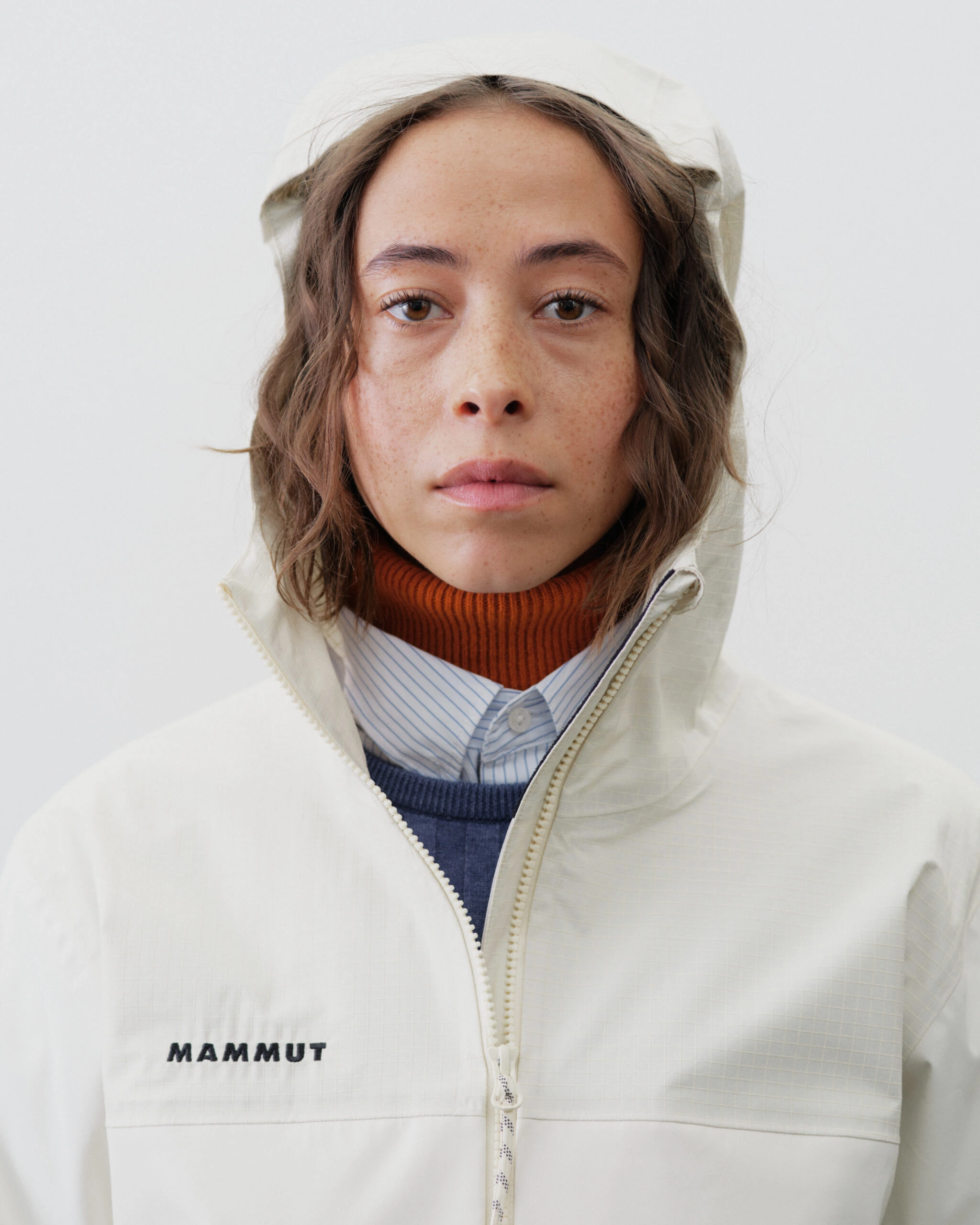 Close-up of a person wearing a cream Mammut hooded jacket with the zipper partially closed, layered over a blue striped shirt and burnt orange turtleneck against a neutral background.